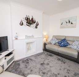 A living room with a TV on a white stand a gray sofa three cushions a lamp and wall decorations at Quayside View in Brixham