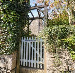 A garden gate leading to a stone house with greenery and a wooden arbor at Westmorland Cottage in Grange-Over-Sands