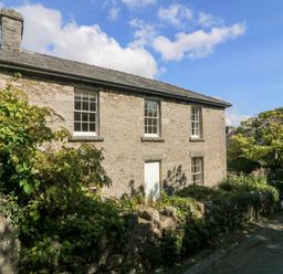 A stone house with white windows and a white door surrounded by greenery next to a road at Westmorland Cottage Grange-Over-Sands