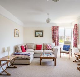 A living room with a beige sofa red and patterned cushions a wooden coffee table armchair with a blue cushion side tables with lamps and red patterned curtains at Thorncroft in West Bay