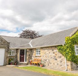 A stone cottage with a single wooden door and white garage door with plants and bench outside at Coquet View Cottage in Rothbury