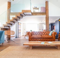 A living room with a leather sofa blue armchair wooden coffee table and a staircase with glass railing at See Two Seas Cape Cornwall in St Just