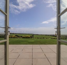 An open patio door leading to a tiled patio and a lawn with fields and a partly cloudy sky at An Gwel in Sennen