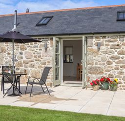 A stone exterior house with an open double door leading inside a patio table with an umbrella and two chairs and flower pots on the ground at An Gwel in Sennen