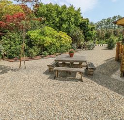 An outdoor area with a wooden picnic table and benches on gravel near a wooden building and surrounded by trees and shrubs at Willow Lodge in St Florence