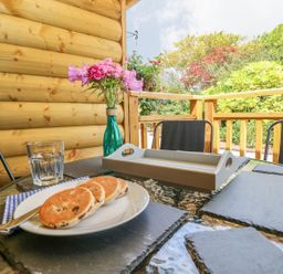 A table with a plate of three scones a glass of water a vase with flowers and patio chairs on a wooden deck outside at Willow Lodge in St Florence