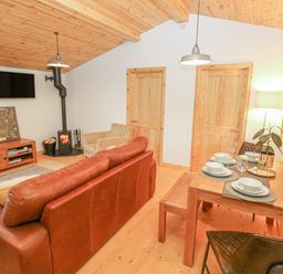 A living room with a brown leather sofa, a wood burning stove, a wall mounted TV, and a dining table set for four at Willow Lodge in St Florence