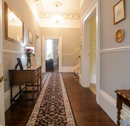 A hallway with wooden floor a patterned runner rug a wooden side table with flowers a mirror and a chandelier at Holcombe House in Torquay