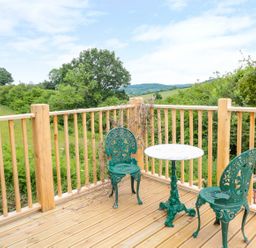 A wooden deck with two green metal chairs and a round table overlooking fields and trees at The Hideout in Llangrove