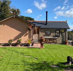 A backyard with a stone and brick house a wood stack flowers in pots a wooden deck and a dog lying on the grass at The Hideout in Llangrove