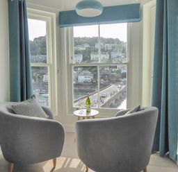 Two grey chairs with cushions and a small round table with a bottle and glasses by a window overlooking a bridge and houses at Harbour View Apartment in Looe