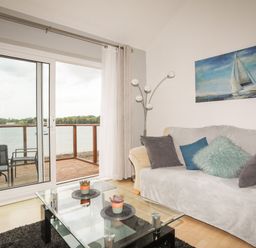 A living room with a sofa pillows a glass coffee table and sliding doors opening to a balcony overlooking water at Delfryd in Y Felinheli