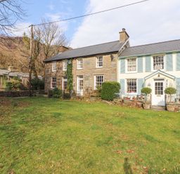 Stone and painted houses with windows and doors facing a lawn with scattered leaves and trees in the background at Brackenbury Cottage in Beddgelert