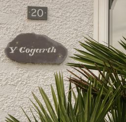 A house number 20 and a name plaque Y Gogarth on a textured wall with green palm leaves in front at Y Gogarth in Benllech