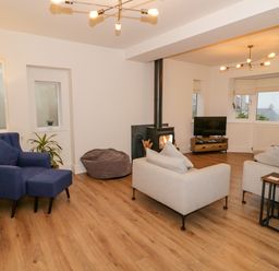 A living room with a blue armchair, beige sofas, a wood stove, a television on a wooden stand, and wooden flooring at Y Gogarth in Benllech