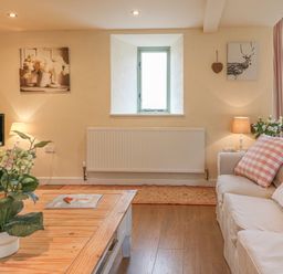 A living room with a beige sofa and pillows a wooden coffee table with a plant a small window and framed pictures at The Apple Barn at Wyke Farm in Lyme Regis