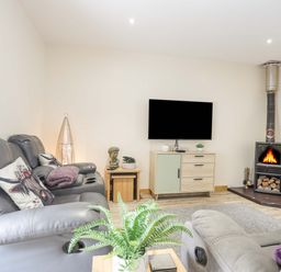 A living room with leather sofas a wooden cabinet a wall-mounted TV and a wood-burning stove at Ffynnon Bach in Deiniolen