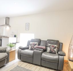 A living room with grey leather sofas and a small kitchen area with a cooker and microwave at Ffynnon Bach in Deiniolen