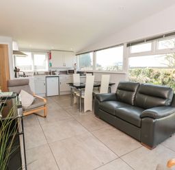 An open living area with a black leather sofa, a dining table with white chairs, a grey armchair, kitchen cabinets, and large windows at 201 Norton Park in Dartmouth