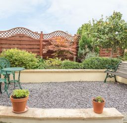 A small garden area with a wooden bench green metal table and chairs potted plants and a variety of shrubs and trees at Church Road Annexe in Cinderford