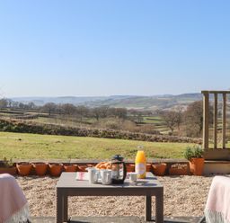 An outdoor patio with a table set with mugs a French press orange juice and croissants overlooking fields and hills at The Shippon at Wyke Farm in Lyme Regis