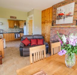 A dining area with a vase of flowers on the table a gray loveseat with red pillows wooden cabinets a brick fireplace and a kitchen with wooden cabinets in the background at Sea View Hideaway in Lahinch County Clare