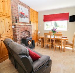 A dining area with a wooden table and chairs next to a window with a red blind and a seating area with grey recliners and a brick fireplace at Sea View Hideaway in Lahinch County Clare