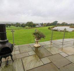 A patio with a chiminea and a planter with flowers overlooking a grassy field and a pond at Nicky Nook Lodge in Scorton
