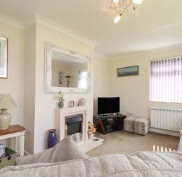 A living room with a TV on a stand a fireplace with decorations a side table with a lamp and magazines and a window with blinds at Tremethic in Trewoon