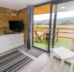 A room with wooden walls a white dresser a television a lamp a striped rug and patio doors leading to a balcony with a table chairs and a hot tub at Cilan Lodge in Llandrillo