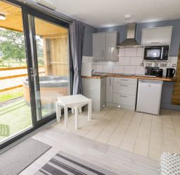 A small kitchen with gray cabinets microwave refrigerator and two white nested tables near sliding glass doors leading to an outdoor area with a hot tub and wooden chairs at Cilan Lodge in Llandrillo