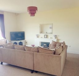 A living room with a beige sofa, a TV on a white stand, a fireplace with a clock and flowers, and a purple lampshade at Llys Madoc The Top Flat in Penmaenmawr