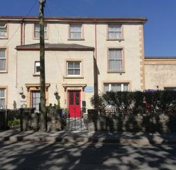 A beige building with multiple windows and a red door with a stone wall and a hedge in front at Llys Madoc The Top Flat in Penmaenmawr