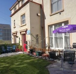An outdoor patio area with chairs tables potted plants and an umbrella outside a building with a red door at Llys Madoc The Top Flat in Penmaenmawr