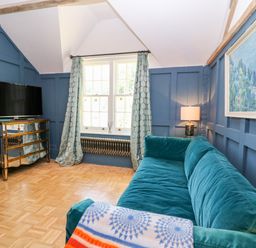 A room with blue paneled walls a teal sofa a window with curtains a television on a mirrored cabinet and a lamp at The Hemmel in Faringdon
