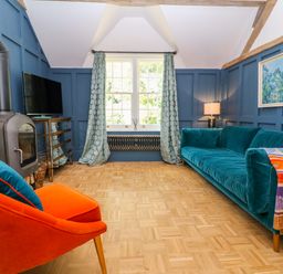 A living room with a blue sofa orange chair wood stove television and window with patterned curtains at The Hemmel in Faringdon