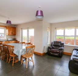 A dining area with wooden chairs around a table and a kitchen with wooden cabinets next to a sitting area with black leather armchair and sofa at Doornogue in Churchtown near Fethard-on-Sea