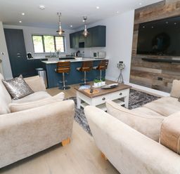 An open living room and kitchen with beige sofas a wooden coffee table a mounted television and a kitchen island with stools at Driftwood Haven in Carbis Bay