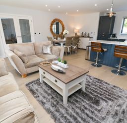 An open living area with beige sofas a center table on a shaggy rug a dining table with wicker chairs and a kitchen island with bar stools at Driftwood Haven in Carbis Bay