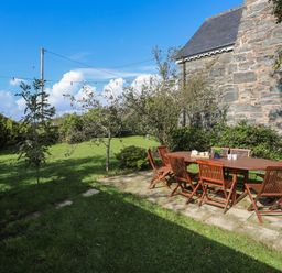 An outdoor patio with wooden table and chairs next to a stone building and grassy lawn at Glyn Morfa in Harlech