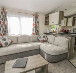 A living room with a beige and gray corner sofa, patterned curtains, a wooden coffee table, and a view of a kitchen area at The Maple in Cloughton