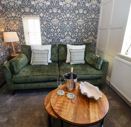 A green sofa with cushions beside a wooden round coffee table holding a champagne bucket and glasses in a living room with floral wallpaper at Gavel Cottage in Whitby