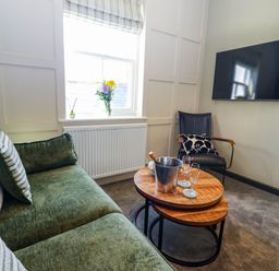 A living room with a green sofa, round wooden coffee tables, a black chair, a vase with flowers on the windowsill, and a wall-mounted television at Gavel Cottage in Whitby