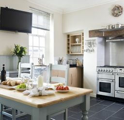 A kitchen with a wooden table set with bread fruit and wine next to a stainless steel stove and a wall mounted television at Albany House Whitby