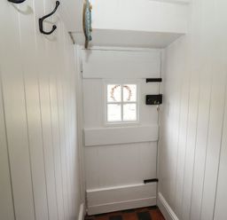 A white wooden door with a small window and black latch in a narrow hallway with white panelled walls at The Cottage in Sandsend