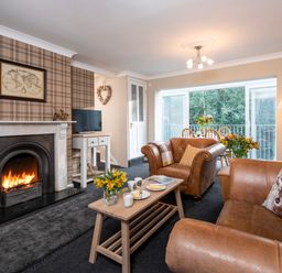 A living room with a fireplace two brown leather armchairs a wooden coffee table and a dining table by large windows at Esk View Cottage in Grosmont