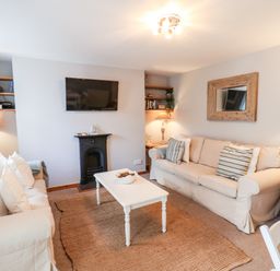 A living room with two beige sofas a white coffee table a wall-mounted TV a small black fireplace wooden shelves and lamps at Albany Cottage in Whitby