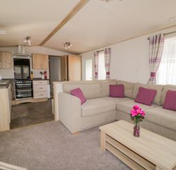 A living room with a beige sectional sofa and purple cushions next to a wooden coffee table with flowers in a vase and an open kitchen with wooden cabinets in the background at The Hideaway in Cockermouth