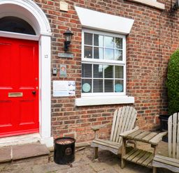 A red door with the number 13 on a brick building next to two wooden chairs and a small wooden table at The Riddle in Whitby
