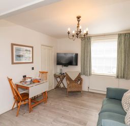 A living room with a dining table and chairs a sofa with cushions a wicker chair a wall mounted TV and a window with curtains at The Riddle in Whitby